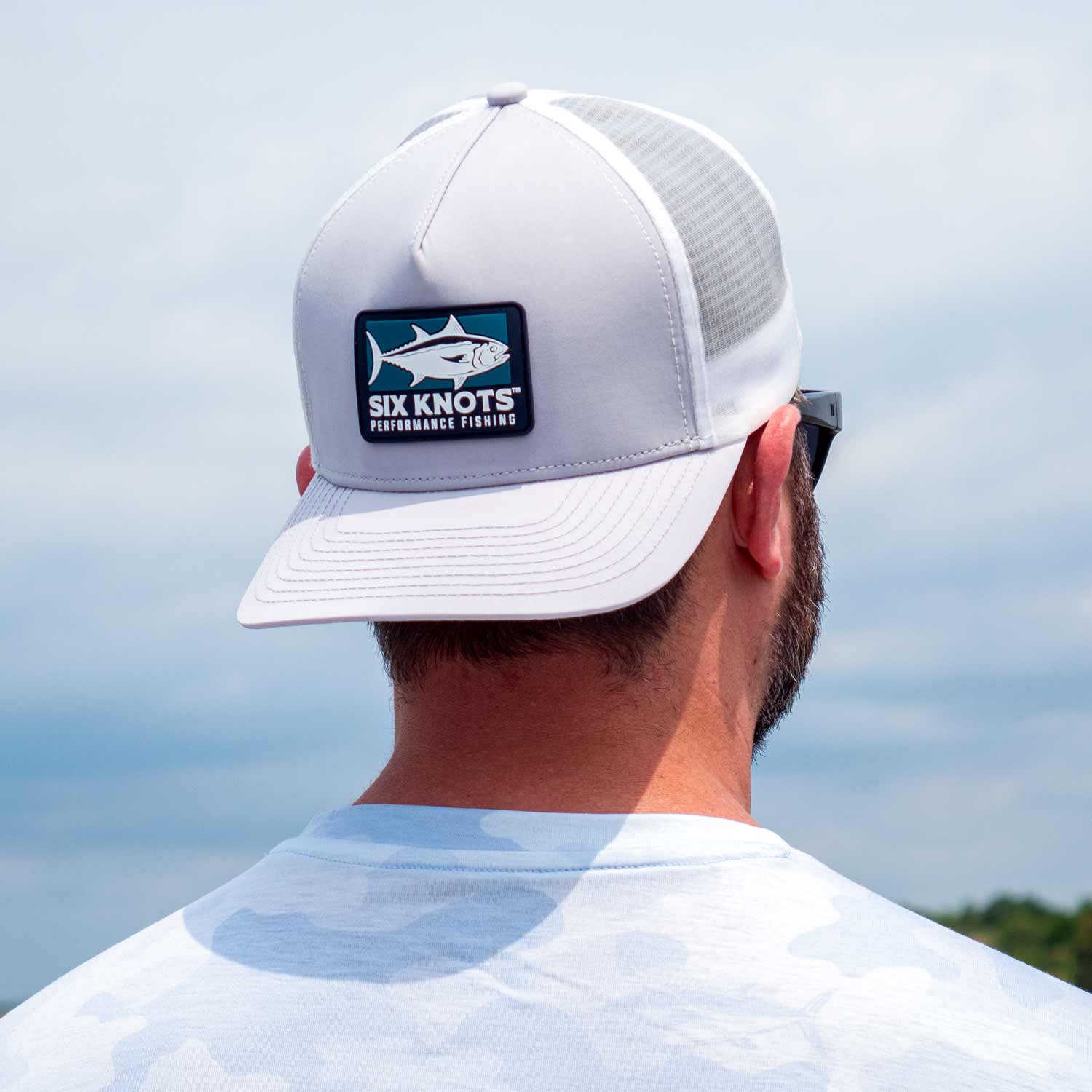 Rear view of man gazing into the distance while wearing the Six Knots Tuna Chaser Snapback Hat backwards. This light gray, high-performance, water-resistant snapback features a structured fit, breathable mesh back, and bold Six Knots Performance Fishing patch logo. Designed for saltwater fishing, boating, and sunny coastal adventures, it offers durable protection and all-day comfort on the water.