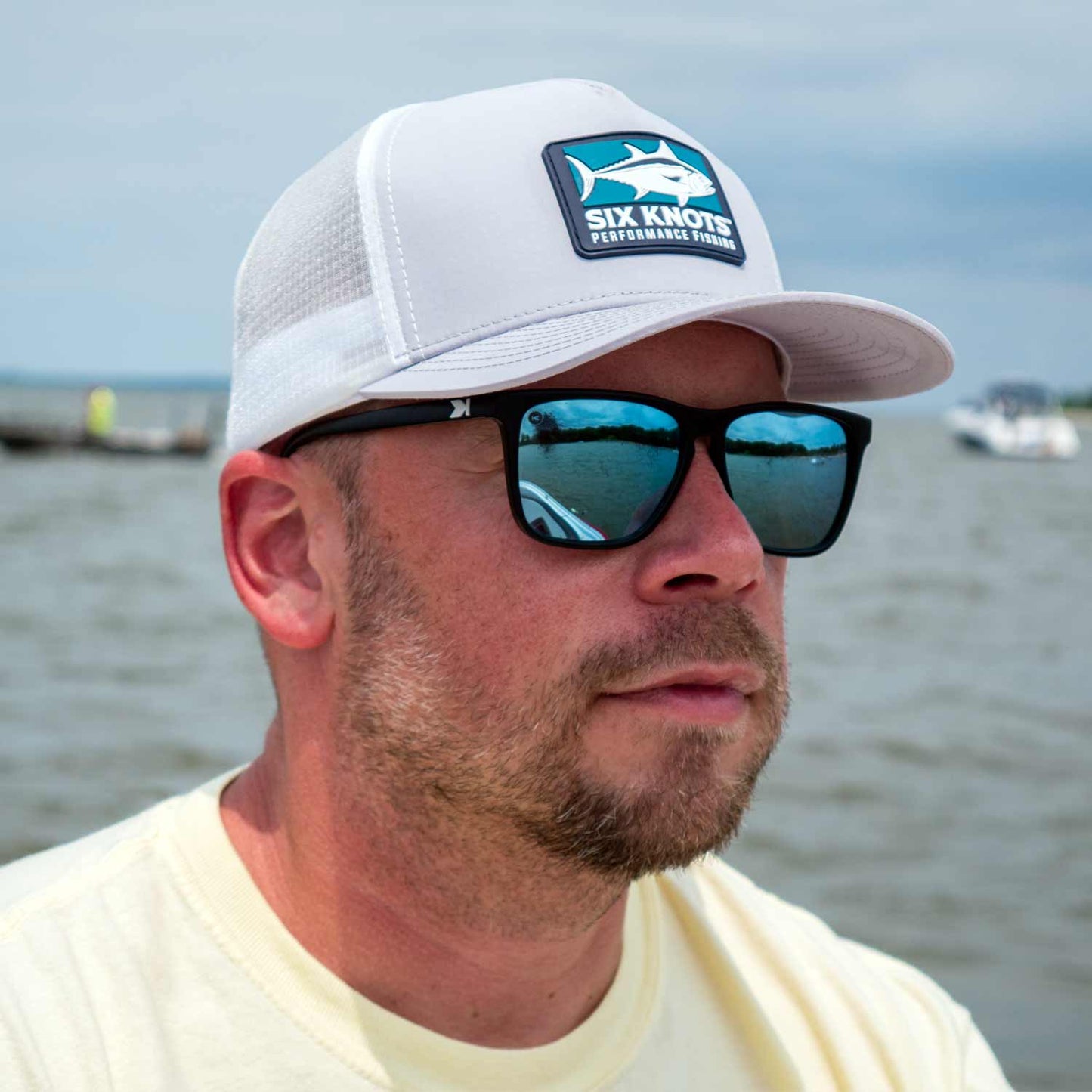 Man on the water gazing into the distance while wearing the Six Knots Tuna Chaser Snapback Hat and polarized sunglasses. This light gray, high-performance, water-resistant snapback features a structured fit, breathable mesh back, and bold Six Knots Performance Fishing patch logo. Designed for saltwater fishing, boating, and sunny coastal adventures, it offers durable protection and all-day comfort on the water.