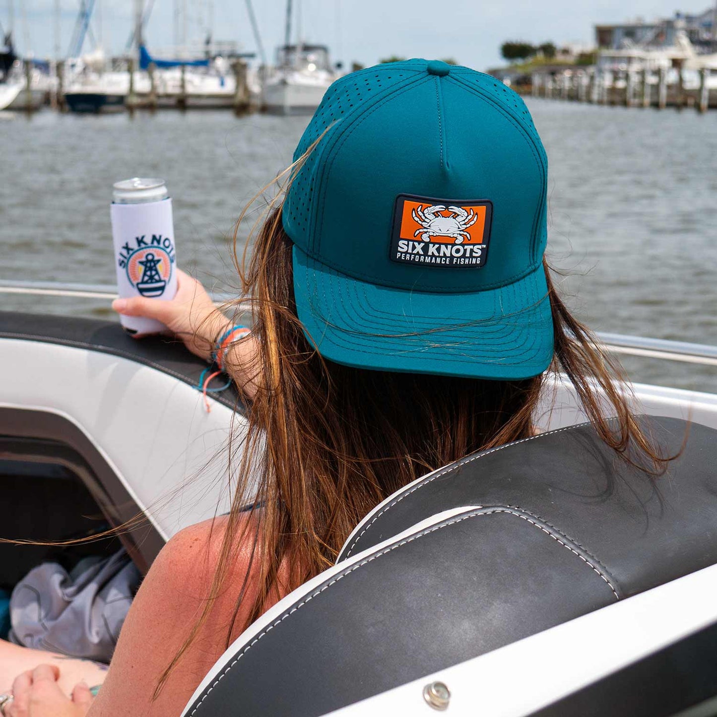 Woman relaxing on a powerboat, holding a canned drink and gazing into the distance while wearing the Six Knots Waterman Snapback Hat backwards. This teal, water-resistant performance snapback features a structured fit, breathable mesh back, and bold Six Knots Performance Fishing patch logo. Engineered for saltwater fishing, offshore boating, beach days, and sunny coastal adventures, it delivers durable sun protection, moisture control, and all-day comfort on the water.