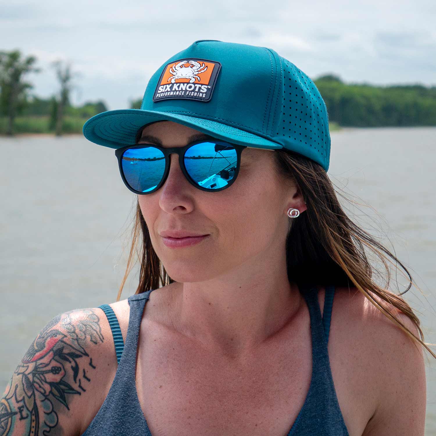 Woman on the water gazing into the distance while wearing the Six Knots Waterman Snapback Hat and polarized sunglasses. This teal, high-performance, water-resistant snapback features a structured fit, breathable mesh back, and bold Six Knots Performance Fishing patch logo. Designed for saltwater fishing, boating, and sunny coastal adventures, it offers durable protection and all-day comfort on the water.