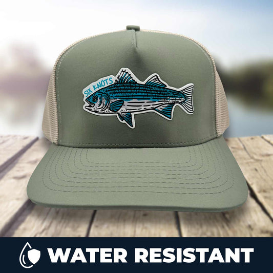 Six Knots Trophy Season Snapback Hat resting on a dock with a blurred marina backdrop — water-resistant performance fishing hat in green and tan, featuring a bold front patch with an embroidered striped bass and Six Knots lettering. Built for durability and breathability, ideal for saltwater fishing, offshore boating, and rugged all-weather coastal lifestyle wear.