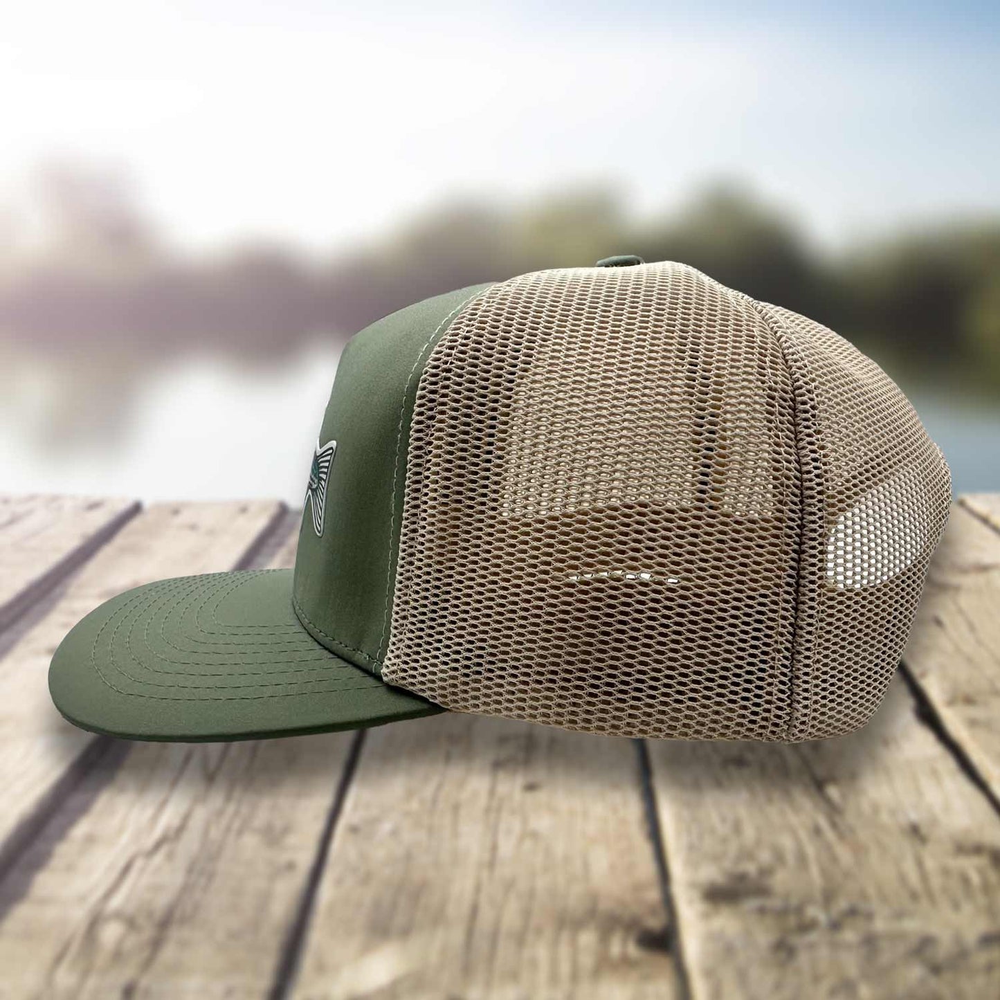 Side view of the Six Knots Trophy Season Snapback Hat resting on a dock with a blurred marina backdrop, showcasing the breathable mesh back — durable, water-resistant fishing hat in green and tan with an embroidered striped bass patch and bold Six Knots branding. Designed for performance and comfort, perfect for saltwater fishing, offshore boating, and all-weather coastal adventures.