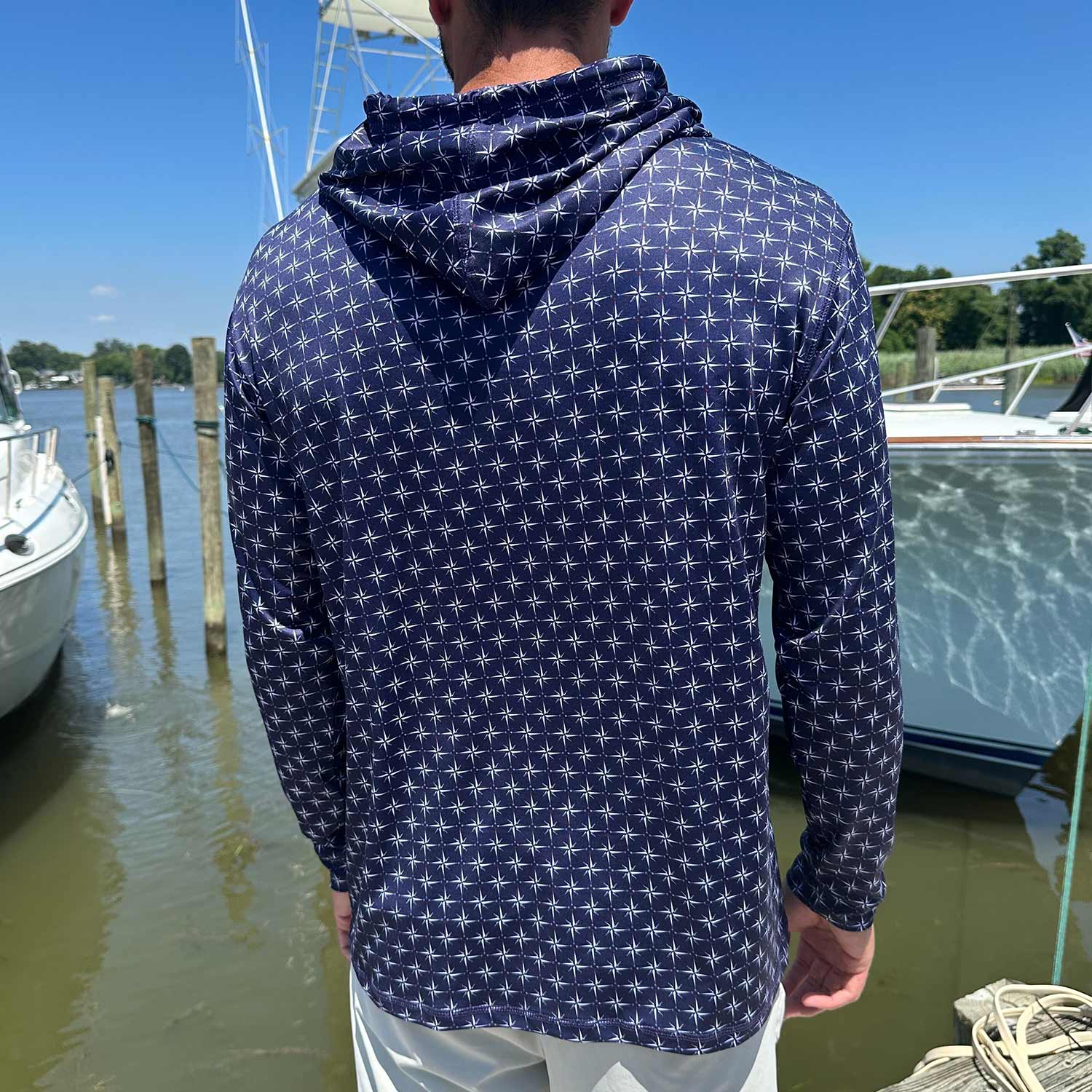 Rear view of man in a marina wearing the Six Knots True North Men's Performance Hoodie in navy with a white all-over compass print. This moisture-wicking, quick-dry hoodie delivers UPF sun protection and breathable, lightweight performance—perfect for boating, saltwater fishing, and all-day coastal wear in warm weather.