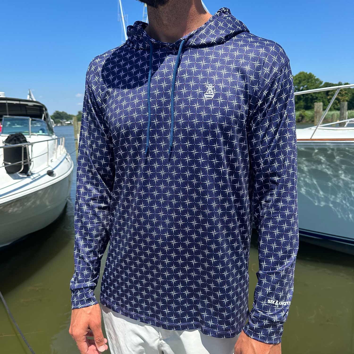 Man standing in a marina wearing the Six Knots True North Men's Performance Hoodie in navy, featuring an all-over white compass pattern pointing true north. This lightweight, moisture-wicking hoodie offers UPF sun protection and breathable comfort—ideal for boating, saltwater fishing, and warm-weather coastal adventures.