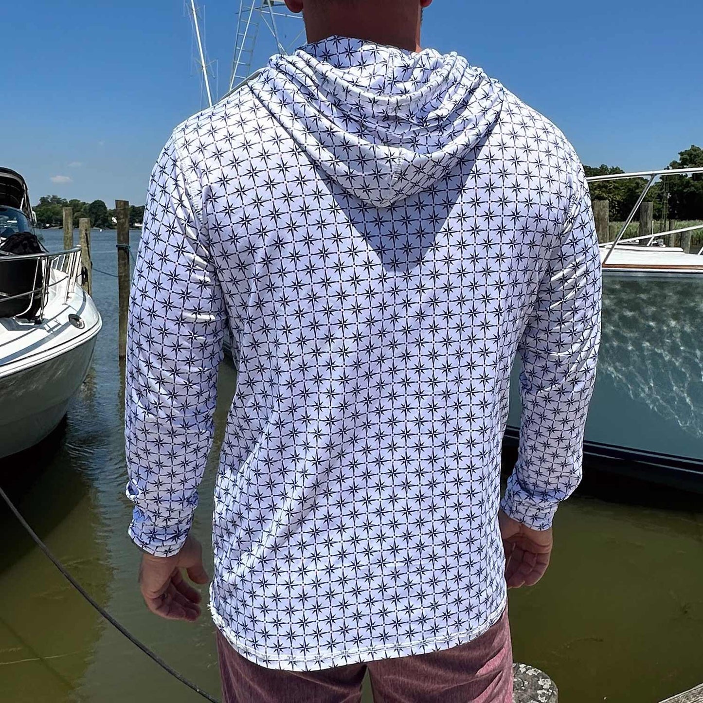 Rear view of man in a marina wearing the Six Knots True North Men's Performance Hoodie in white with a navy blue all-over compass print. This moisture-wicking, quick-dry hoodie delivers UPF sun protection and breathable, lightweight performance—perfect for boating, saltwater fishing, and all-day coastal wear in warm weather.