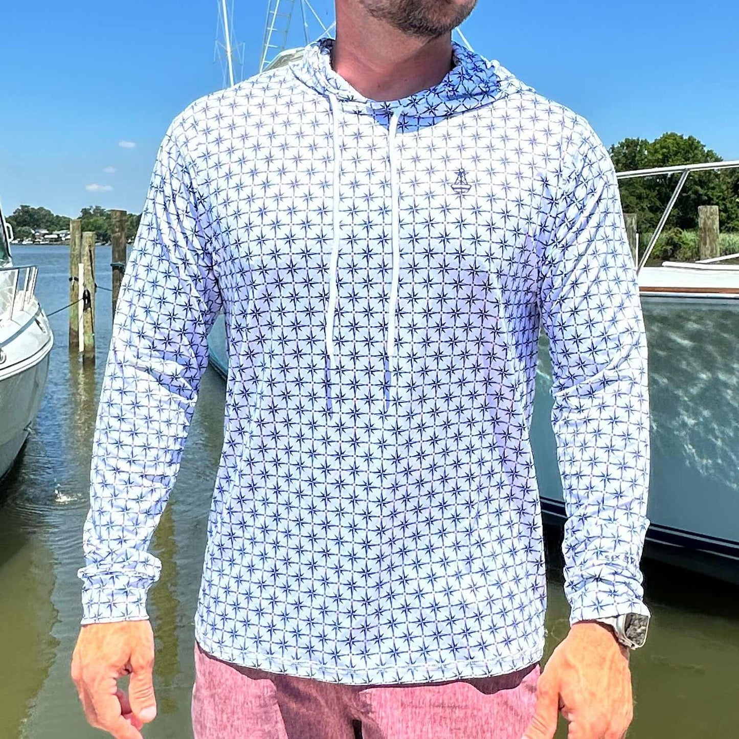 Man standing in a marina wearing the Six Knots True North Men's Performance Hoodie in white, featuring an all-over navy blue compass pattern pointing true north. This lightweight, moisture-wicking hoodie offers UPF sun protection and breathable comfort—ideal for boating, saltwater fishing, and warm-weather coastal adventures.