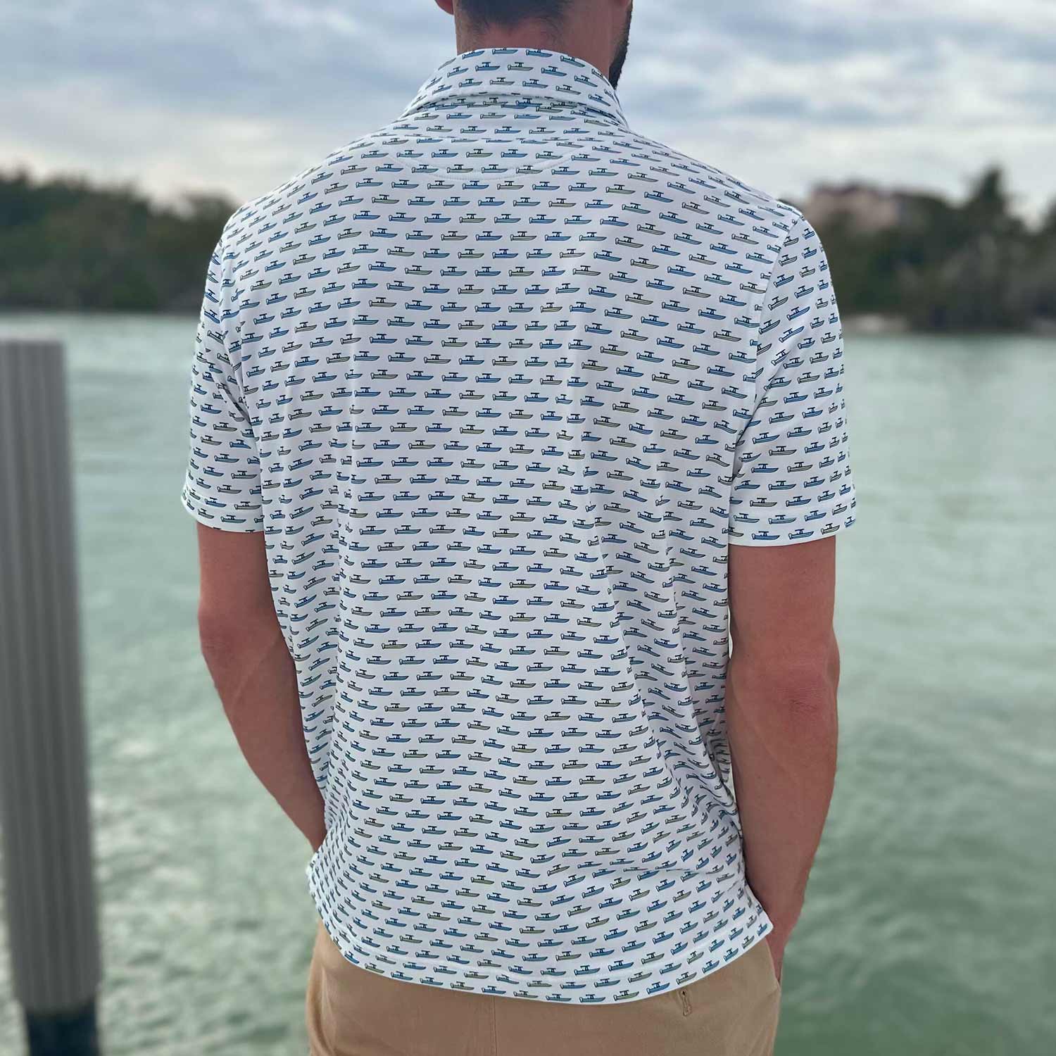 Rear view of man standing on a dock in Florida wearing the Six Knots Center Console Performance Polo Shirt. This white men's moisture-wicking polo is lightweight, breathable, and features a vibrant all-over print of green and blue center console fishing boats. Ideal for boating, offshore fishing, golf, beach outings, and everyday coastal lifestyle apparel.