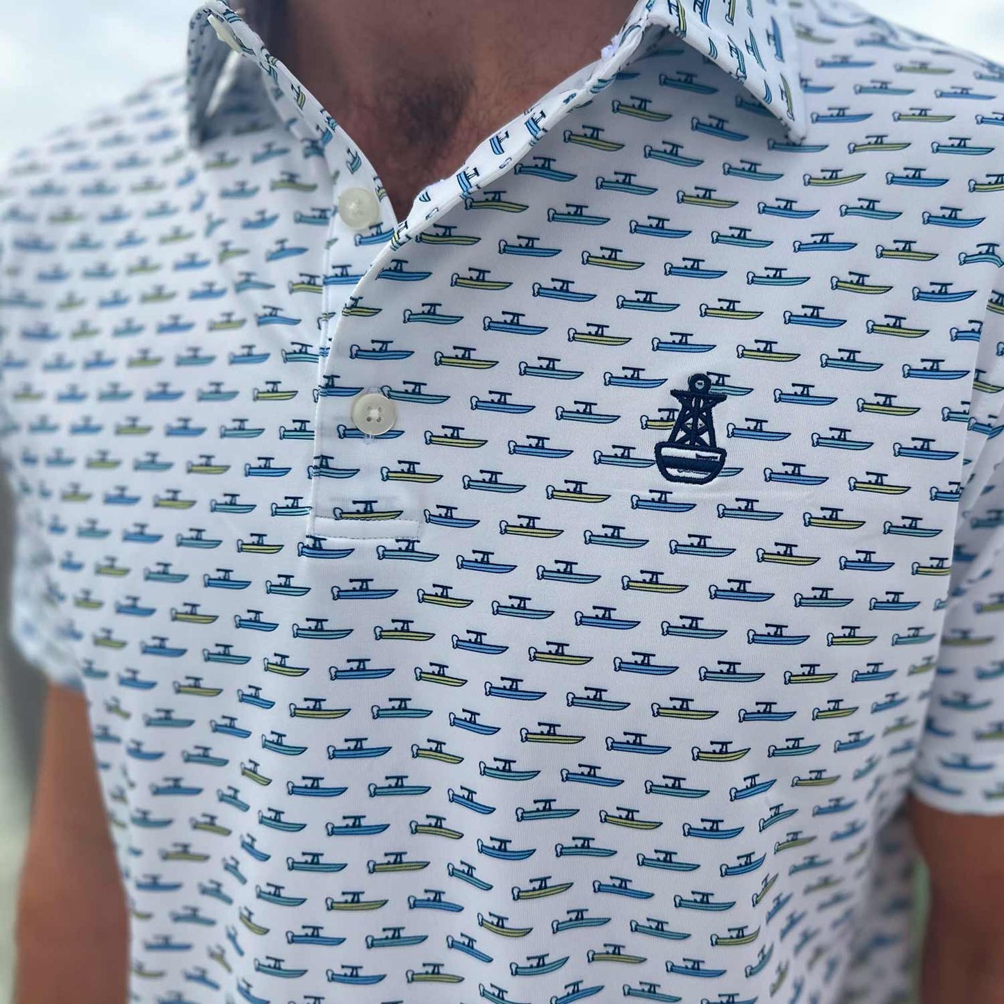 Close-up detail of the Six Knots Center Console Performance Polo Shirt featuring the signature Six Knots buoy logo. This white men's performance polo is lightweight, breathable, and made with moisture-wicking fabric—showcasing a vibrant all-over print of green and blue center console fishing boats. Perfect for boating, saltwater fishing, golf, beach trips, and everyday coastal lifestyle wear.