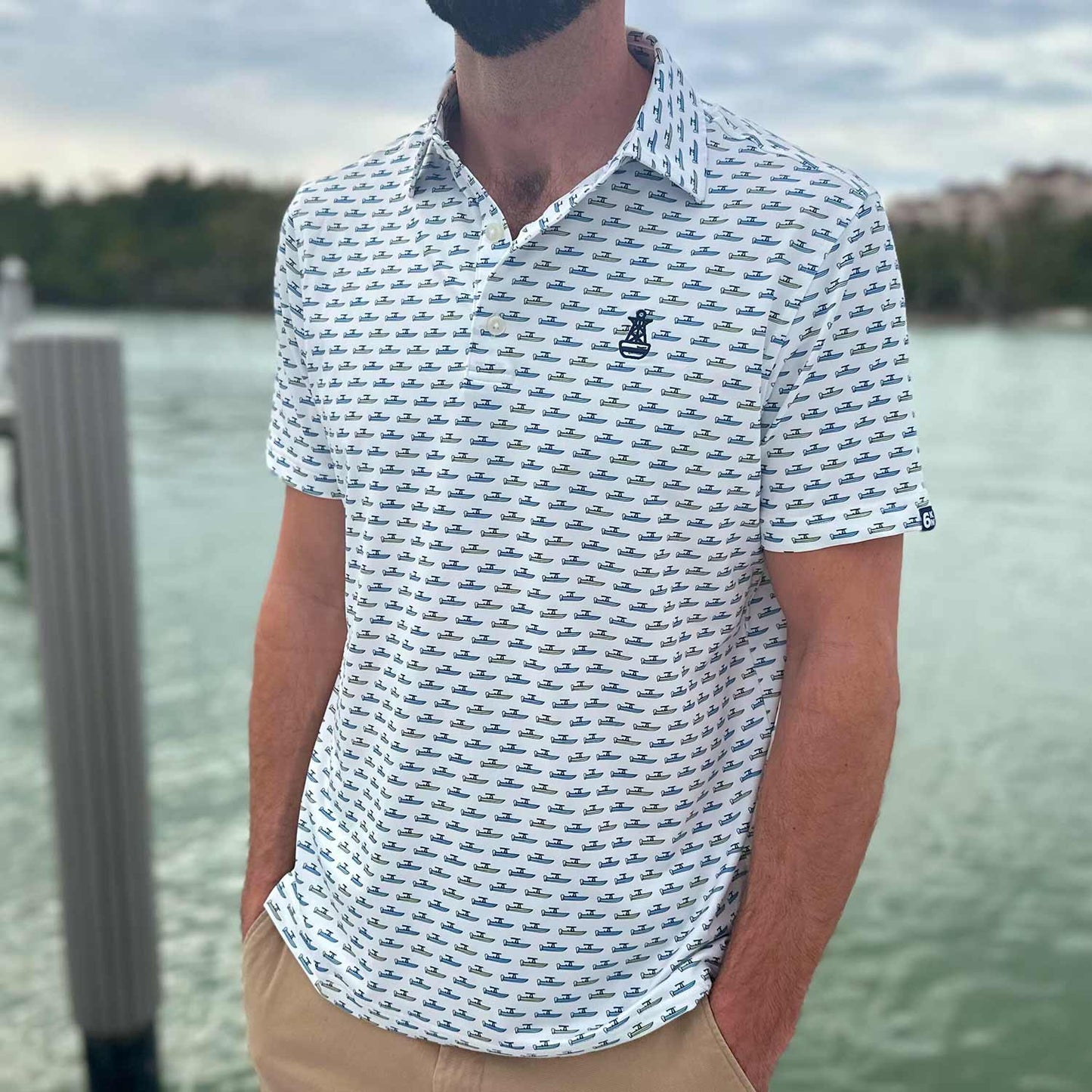 Man standing on a dock in Florida wearing the Six Knots Center Console Performance Polo Shirt. This white men's performance polo is lightweight, breathable, and made with moisture-wicking fabric—featuring a vibrant all-over print of green and blue center console boats. Perfect for boating, fishing, golf, beach trips, and everyday coastal lifestyle wear.