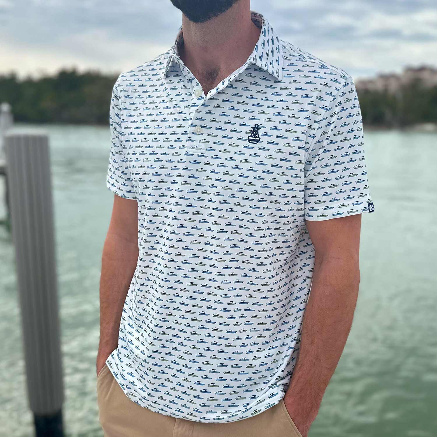 Man standing on a dock in Florida wearing the Six Knots Center Console Performance Polo Shirt. This white men's performance polo is lightweight, breathable, and made with moisture-wicking fabric—featuring a vibrant all-over print of green and blue center console boats. Perfect for boating, fishing, golf, beach trips, and everyday coastal lifestyle wear.