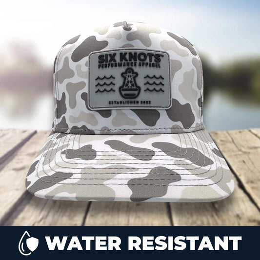 Six Knots Coastal Camo Snapback Hat resting on a dock with a blurred marina backdrop — stylish, water-resistant hat featuring a gray camouflage pattern and a front patch reading ‘Six Knots Performance Apparel Established 2022’; perfect for fishing, boating, and all-weather coastal wear.