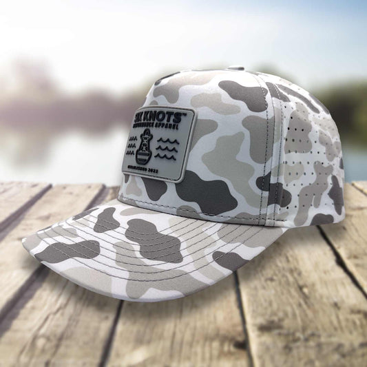 Angled side view of the Six Knots Coastal Camo Snapback Hat on a dock with a blurred marina background — water-resistant performance hat with a gray camo pattern and embroidered ‘Six Knots Performance Apparel Established 2022’ patch; ideal for fishing, boating, and everyday coastal lifestyle wear.