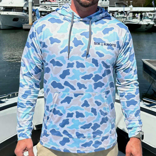 Man standing on a boat in a marina wearing the Six Knots Coastal Camo Performance Hoodie in the Marsh colorway. This lightweight, breathable men's fishing hoodie features moisture-wicking fabric and a blue-and-tan coastal camouflage print with contrasting light and dark tones. Ideal for boating, saltwater fishing, outdoor excursions, and versatile all-weather coastal wear.