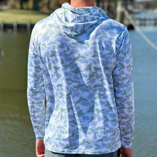Rear view of man in a marina wearing the Six Knots Coastal Camo Performance Hoodie in the ‘Wetlands’ colorway — lightweight, breathable, and quick-dry fishing hoodie featuring a blue and green coastal wetlands camo pattern; UPF sun-protective apparel designed for saltwater fishing, offshore boating, and rugged all-weather coastal adventures.