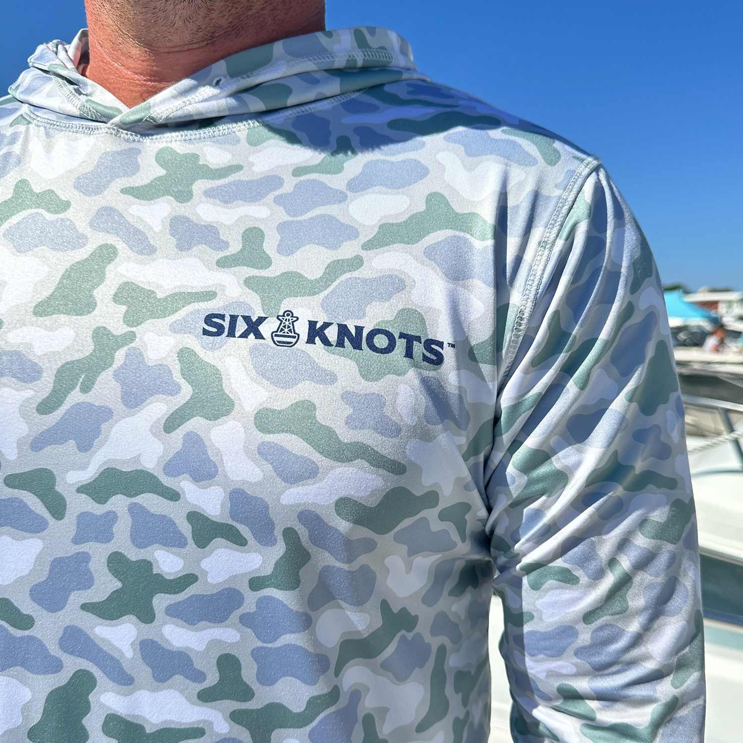 Close-up detail of the Six Knots Coastal Camo Performance Hoodie in the ‘Wetlands’ colorway, showcasing the classic Six Knots wordmark — lightweight, breathable, quick-dry fishing hoodie with a blue and green camouflage pattern inspired by coastal wetlands; UPF-rated sun protection, ideal for saltwater fishing, offshore boating, and all-weather marine adventures.
