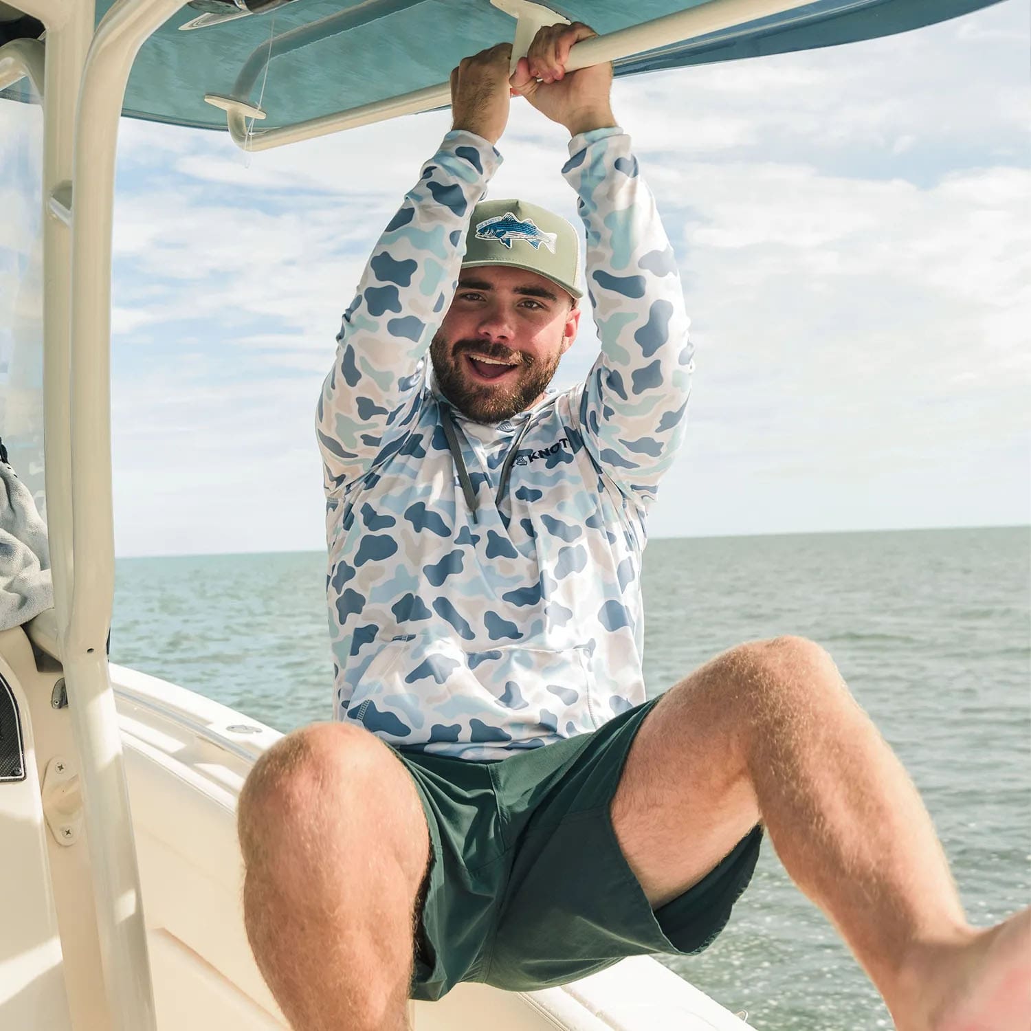 Man leaning off the side of a boat in Fort Lauderdale, Florida wearing a Six Knots Trophy Season water-resistant snapback hat and Coastal Camo Performance Hoodie in Marsh pattern — lightweight boating and fishing apparel