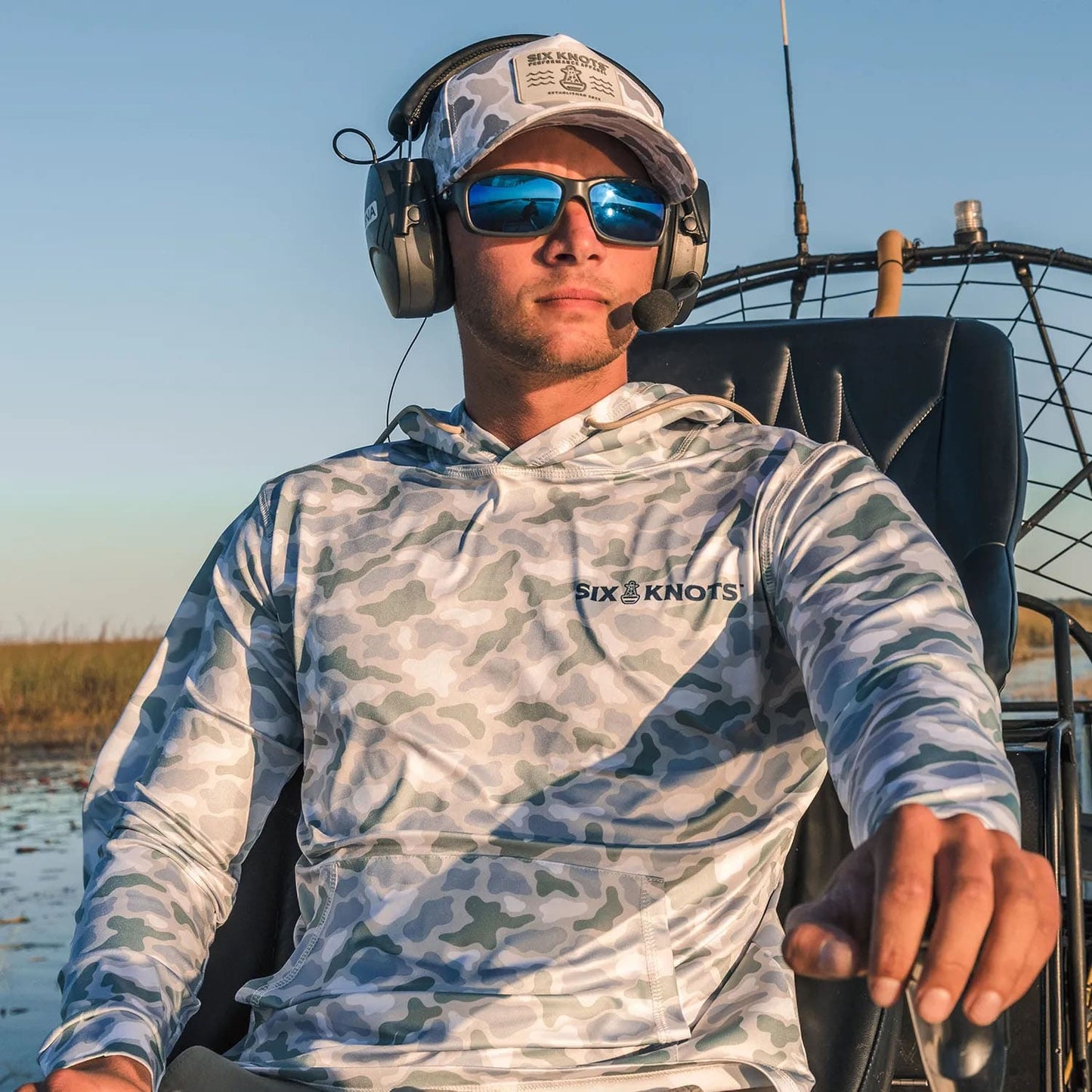 Man airboating in sunglasses, wearing the Six Knots water-resistant Coastal Camo Snapback Hat and Wetlands-pattern Chesapeake Performance Hoodie — lightweight, moisture-wicking boating apparel