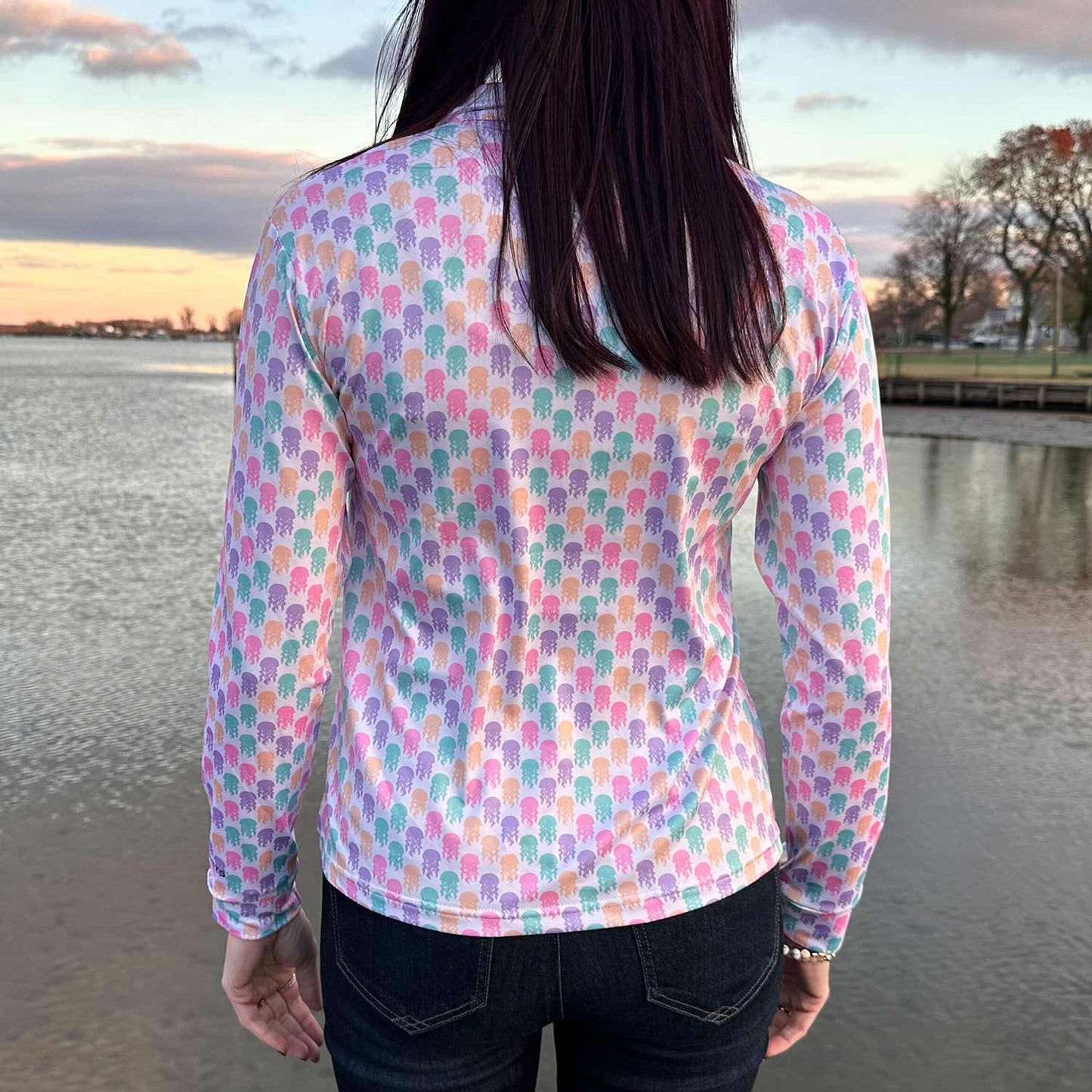 Rear view of woman on a dock at sunset wearing the Six Knots Don't Be Jelly Women's Performance Quarter Zip. This breathable, lightweight women's pullover features moisture-wicking fabric and a vibrant all-over jellyfish print in bold coastal colors — ideal for boating, fishing, beach outings, and active seaside adventures.