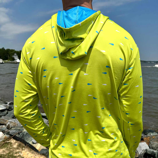 Rear view of man by coastal rocks and ocean wearing the vibrant yellow Six Knots Dorado Performance Fishing Hoodie. This long-sleeve, lightweight hoodie features a bold white and blue dorado fish pattern, breathable moisture-wicking fabric, and built-in UPF 50+ sun protection. Perfect for saltwater fishing, offshore boating, beach trips, and high-performance coastal wear.