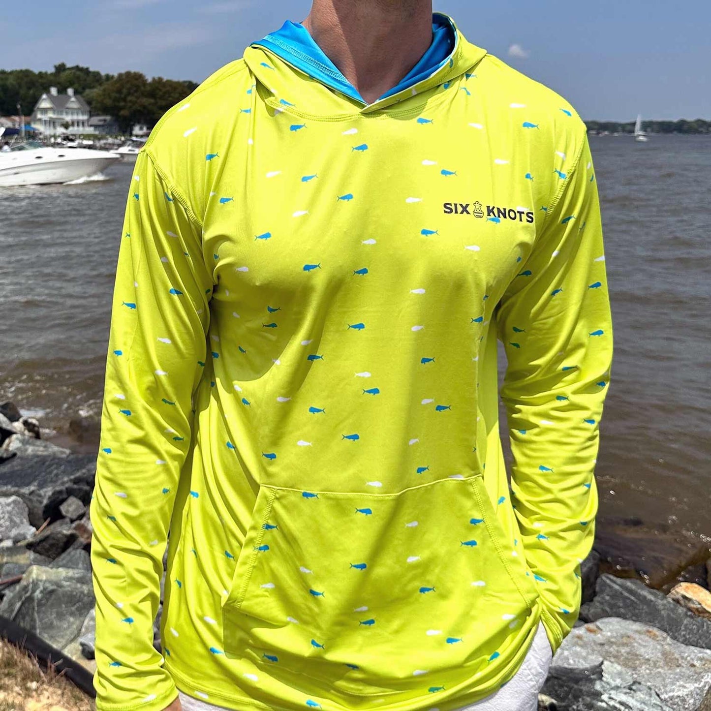 Man standing by coastal rocks and water wearing the vibrant yellow Six Knots Dorado Performance Hoodie. This lightweight, moisture-wicking fishing hoodie features a bold white and blue dorado fish pattern, UPF sun protection, and breathable fabric. Ideal for offshore fishing, boating, beach outings, and active coastal adventures.