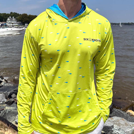Man standing by coastal rocks and water wearing the vibrant yellow Six Knots Dorado Performance Hoodie. This lightweight, moisture-wicking fishing hoodie features a bold white and blue dorado fish pattern, UPF sun protection, and breathable fabric. Ideal for offshore fishing, boating, beach outings, and active coastal adventures.