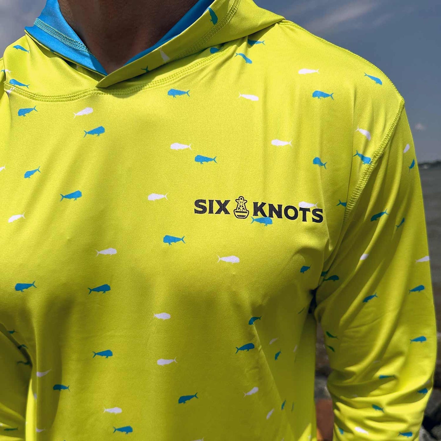 Close-up detail of the vibrant yellow Six Knots Dorado Performance Fishing Hoodie, showcasing a subtle Six Knots wordmark on the chest. This lightweight, long-sleeve fishing hoodie features a striking white and blue dorado fish pattern, breathable moisture-wicking fabric, and UPF 50+ sun protection. Engineered for comfort and durability, it's ideal for offshore fishing, saltwater boating, beach outings, and active coastal adventures.