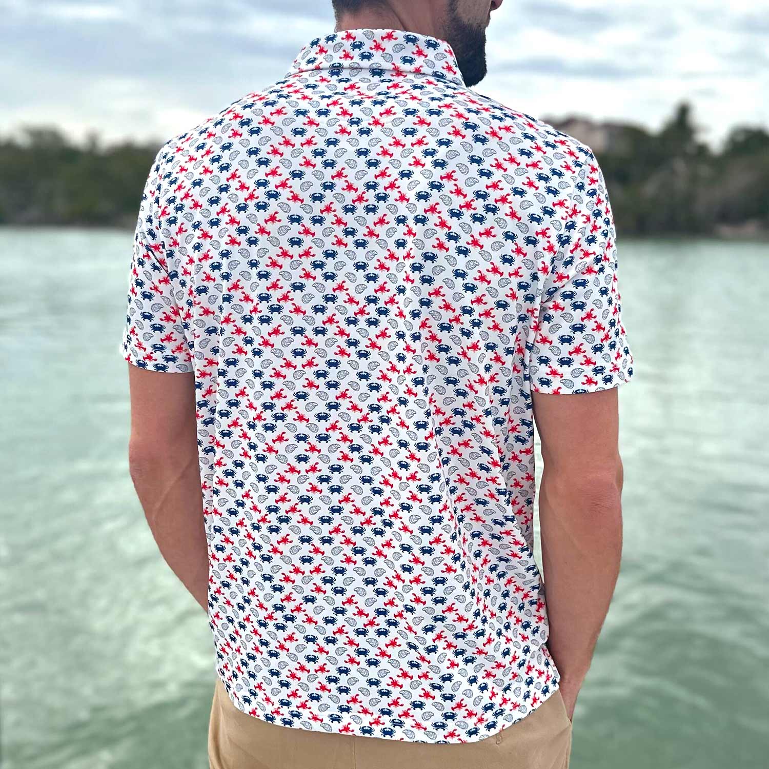 Rear view of a man on a Florida boat dock wearing the Six Knots East Coaster Performance Polo Shirt. This breathable, lightweight men's polo features a red, white, and blue all-over seafood print with iconic East Coast favorites like lobsters, crabs, and oysters. Designed for moisture-wicking comfort, it's ideal for boating, fishing, golf, beach outings, and coastal lifestyle wear