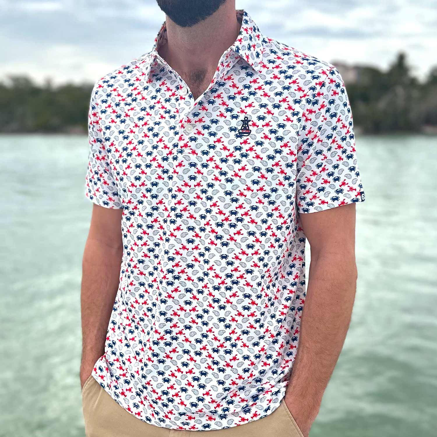 Man standing on a boat dock in Florida wearing the Six Knots East Coaster Performance Polo Shirt. This lightweight, moisture-wicking men's polo features a red, white, and blue all-over seafood print with East Coast favorites like lobsters, crabs, and oysters. Perfect for boating, fishing, golfing, beach trips, and everyday coastal lifestyle wear