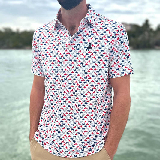 Man standing on a boat dock in Florida wearing the Six Knots East Coaster Performance Polo Shirt. This lightweight, moisture-wicking men's polo features a red, white, and blue all-over seafood print with East Coast favorites like lobsters, crabs, and oysters. Perfect for boating, fishing, golfing, beach trips, and everyday coastal lifestyle wear