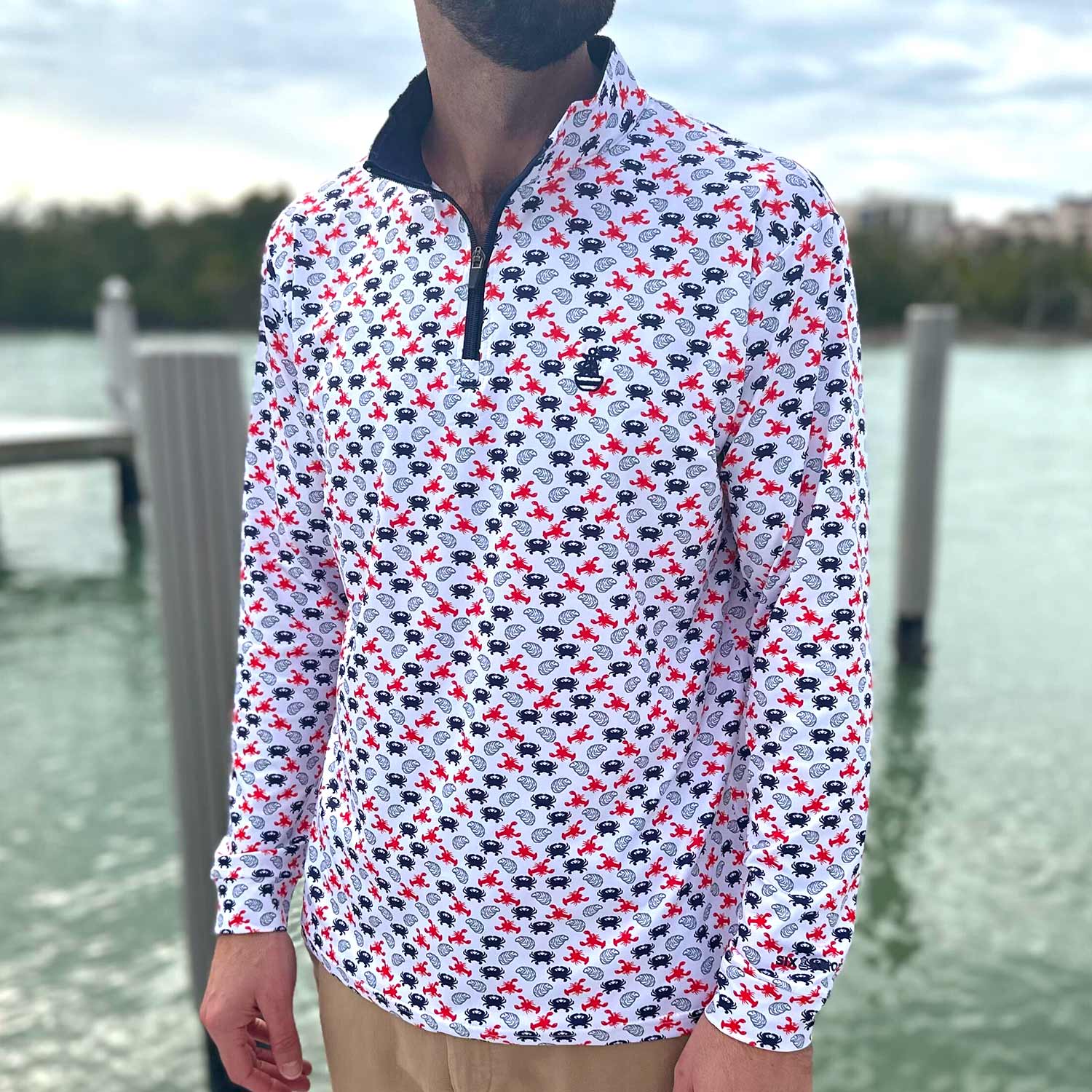 Man standing on a boat dock in Florida wearing the Six Knots East Coaster Performance Quarter Zip — lightweight, moisture-wicking pullover featuring a red, white, and blue all-over print of East Coast seafood favorites like lobsters, crabs, and oysters; ideal for boating, fishing, and coastal lifestyle wear