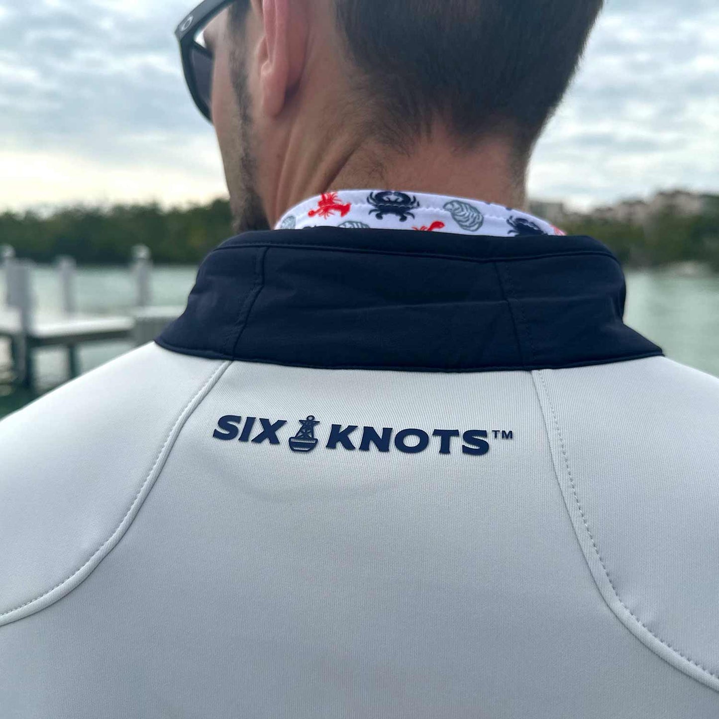 Close-up back detail of the Six Knots Every Day Performance Vest highlighting its collar and navy Six Knots wordmark. This lightweight, breathable men's performance vest features a stylish navy and cream design, providing versatile layering and all-day comfort. Ideal for boating, fishing, outdoor adventures, and everyday coastal lifestyle wear