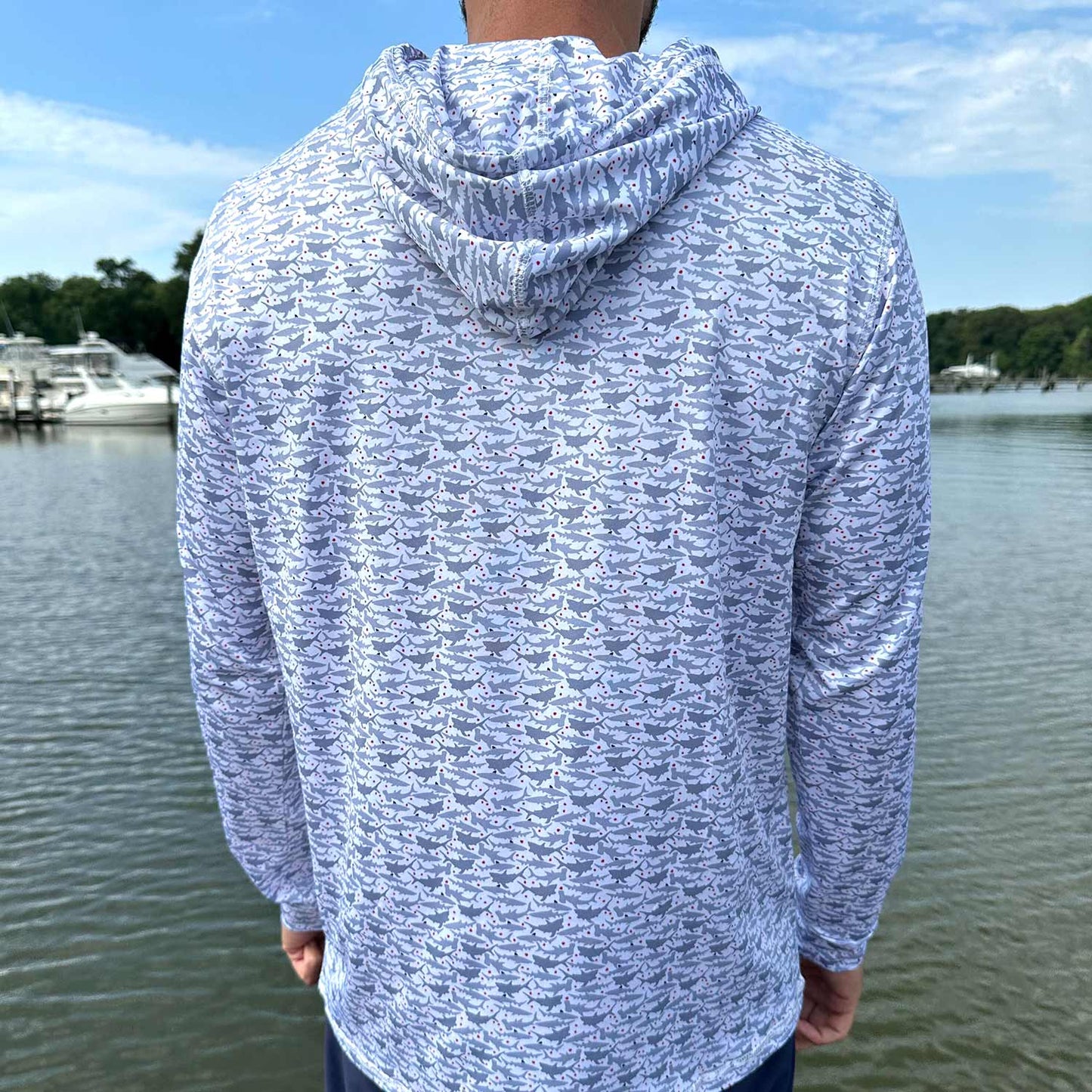 Rear view of man standing by the water wearing the Six Knots Feeding Frenzy Performance Hoodie — lightweight, breathable, and moisture-wicking fishing hoodie with a bold all-over print of feeding sharks and red dots mimicking blood in the water. Designed for UPF sun protection and all-day comfort, perfect for saltwater fishing, offshore boating, and high-performance coastal adventures.