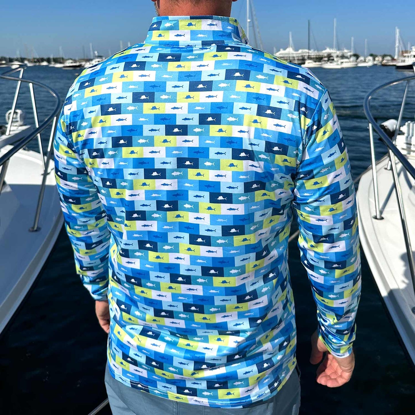 Rear view of man in a marina surrounded by powerboats wearing the Six Knots Fish Flag Men’s Performance Quarter Zip — lightweight, breathable, and moisture-wicking men’s fishing pullover featuring a bold all-over print of sportfish framed in nautical flag-style boxes of lime green, navy, light blue, and white; designed for saltwater fishing, offshore boating, and rugged coastal adventures in any weather.