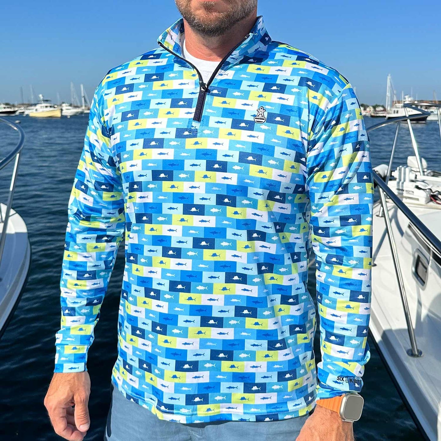 Man standing in a marina surrounded by powerboats wearing the Six Knots Fish Flag Men’s Performance Quarter Zip — lightweight, breathable, and moisture-wicking fishing pullover featuring an all-over fish pattern framed in lime green, navy blue, light blue, and white flag-style boxes; ideal for saltwater angling, offshore boating, and versatile all-weather coastal adventures.