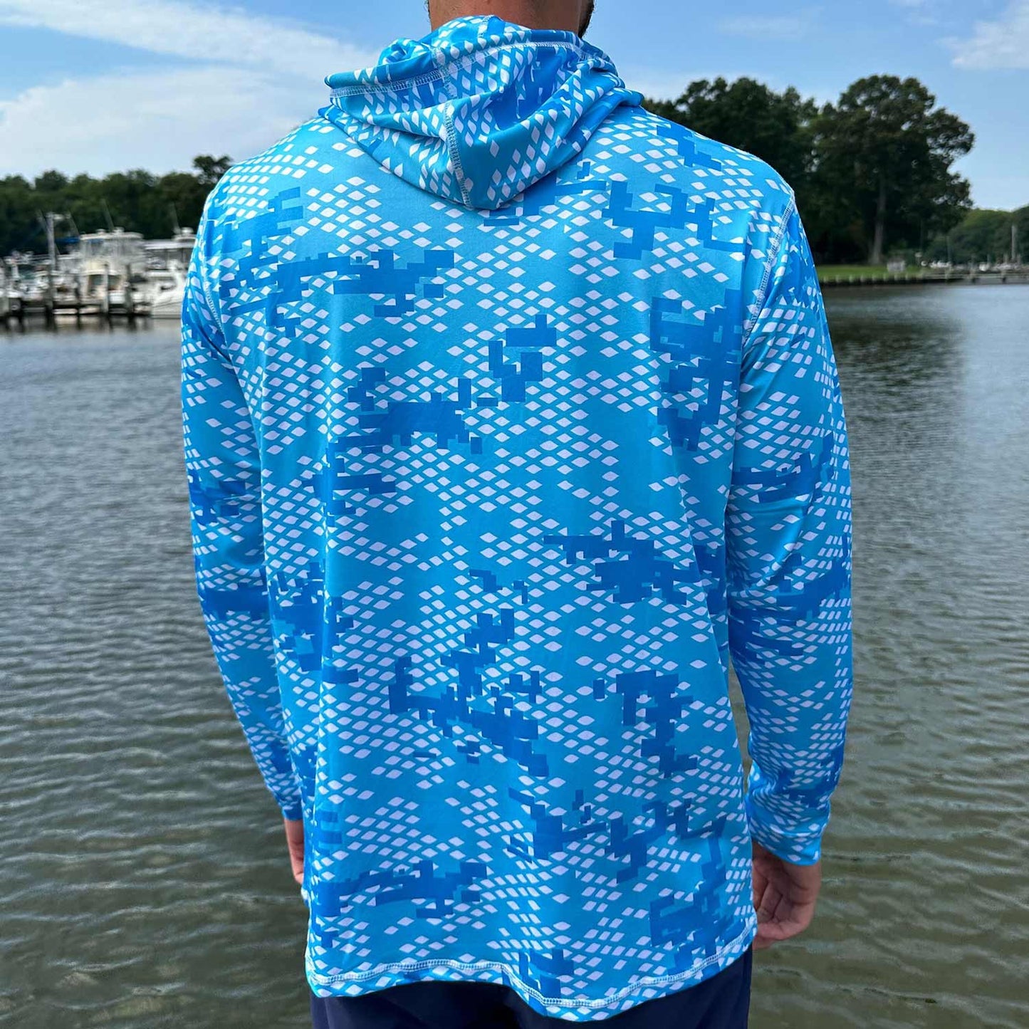 Rear view of man standing near the water wearing the Six Knots Fish Scale Camo Performance Hoodie in blue — performance fishing hoodie featuring a digital fish scale camouflage pattern in blue; lightweight, breathable, and moisture-wicking with UPF sun protection; ideal for saltwater angling, offshore boating, and all-weather coastal adventures.
