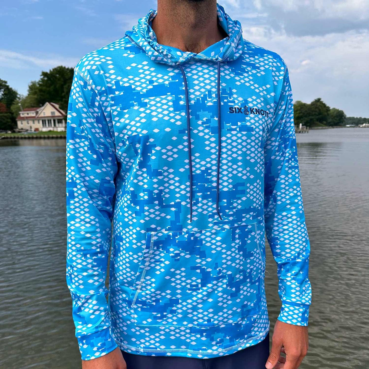 Man standing near the water wearing the Six Knots Fish Scale Camo Performance Hoodie in blue — lightweight, breathable, and moisture-wicking fishing hoodie with a digital blue camo design inspired by fish scales; perfect for saltwater fishing, offshore boating, and rugged coastal adventures in any weather.