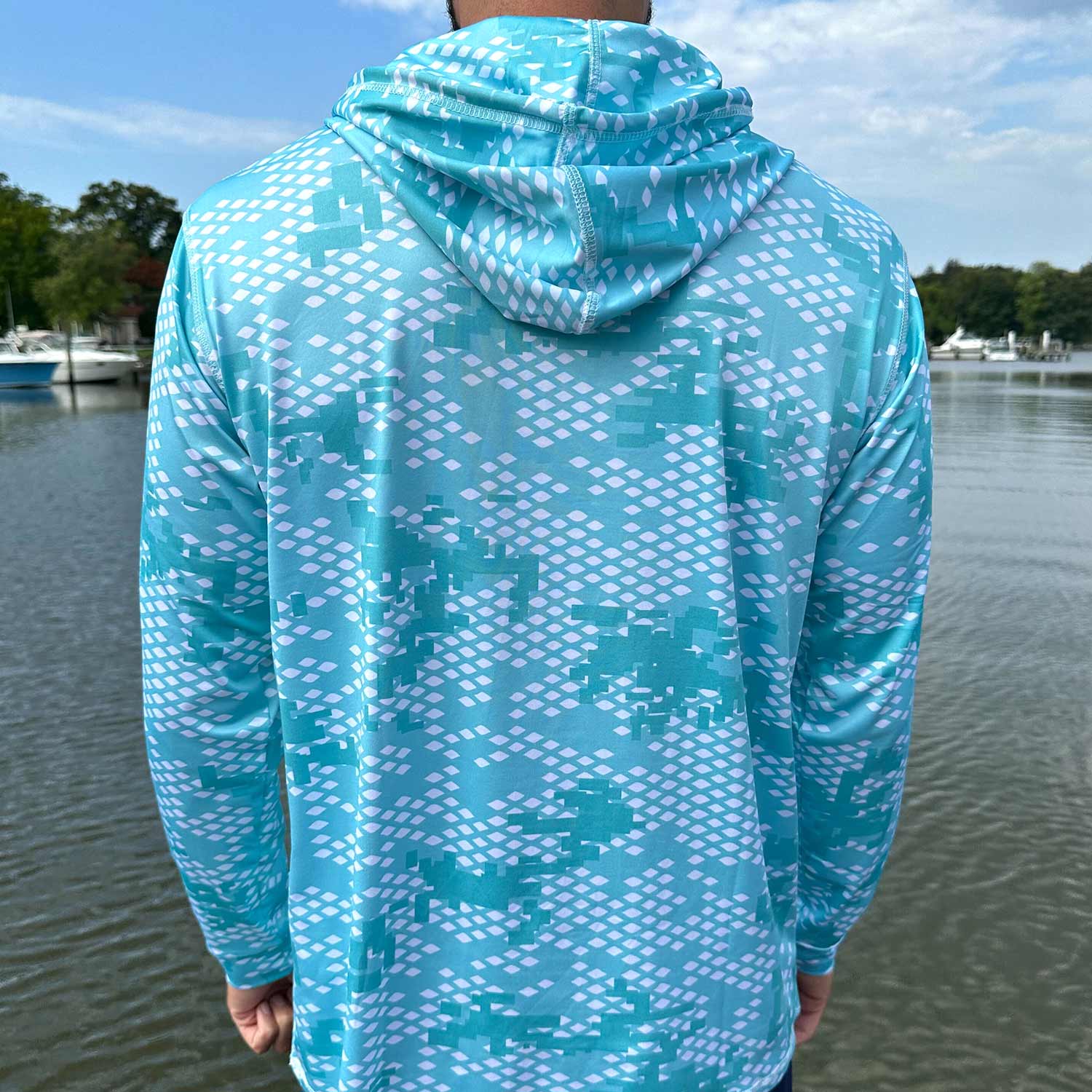 Rear view of man standing near the water wearing the Six Knots Fish Scale Camo Performance Hoodie in green — performance fishing hoodie featuring a digital fish scale camouflage pattern in green; lightweight, breathable, and moisture-wicking with UPF sun protection; ideal for saltwater angling, offshore boating, and all-weather coastal adventures.
