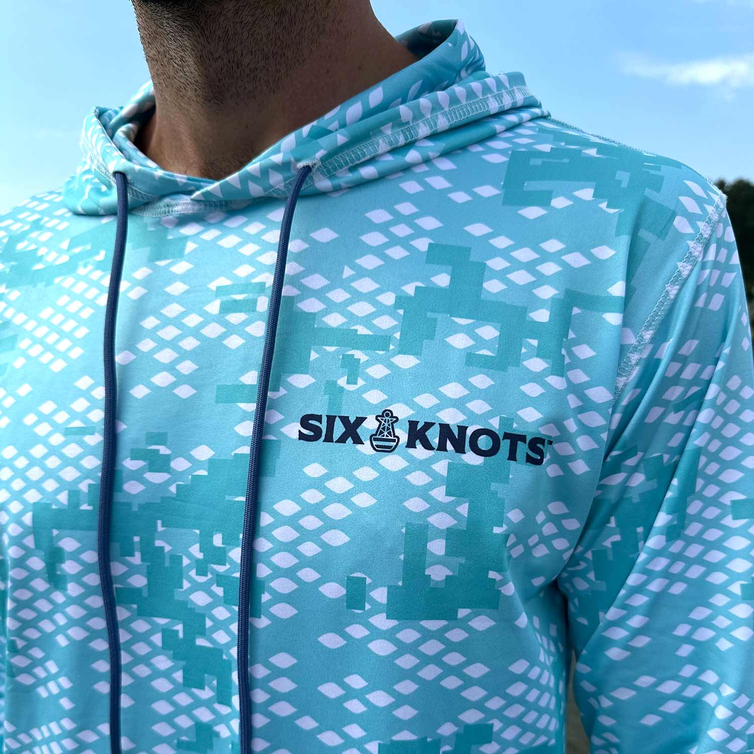 Close-up detail of the Six Knots Fish Scale Camo Performance Hoodie in green, featuring a bold digital camouflage pattern inspired by fish scales and the classic Six Knots wordmark — a high-performance fishing hoodie that’s lightweight, breathable, moisture-wicking, and UPF sun-protective; built for saltwater fishing, offshore boating, and rugged coastal adventures in any weather.