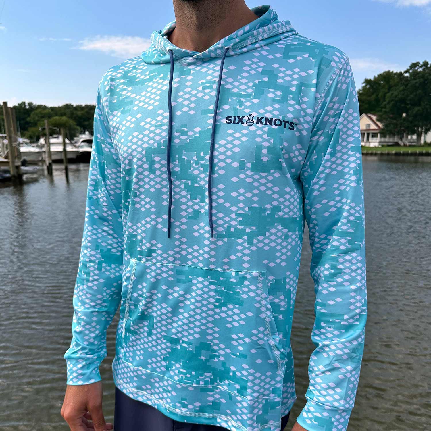 Man standing near the water wearing the Six Knots Fish Scale Camo Performance Hoodie in green — lightweight, breathable, and moisture-wicking fishing hoodie with a digital green camo design inspired by fish scales; perfect for saltwater fishing, offshore boating, and rugged coastal adventures in any weather.