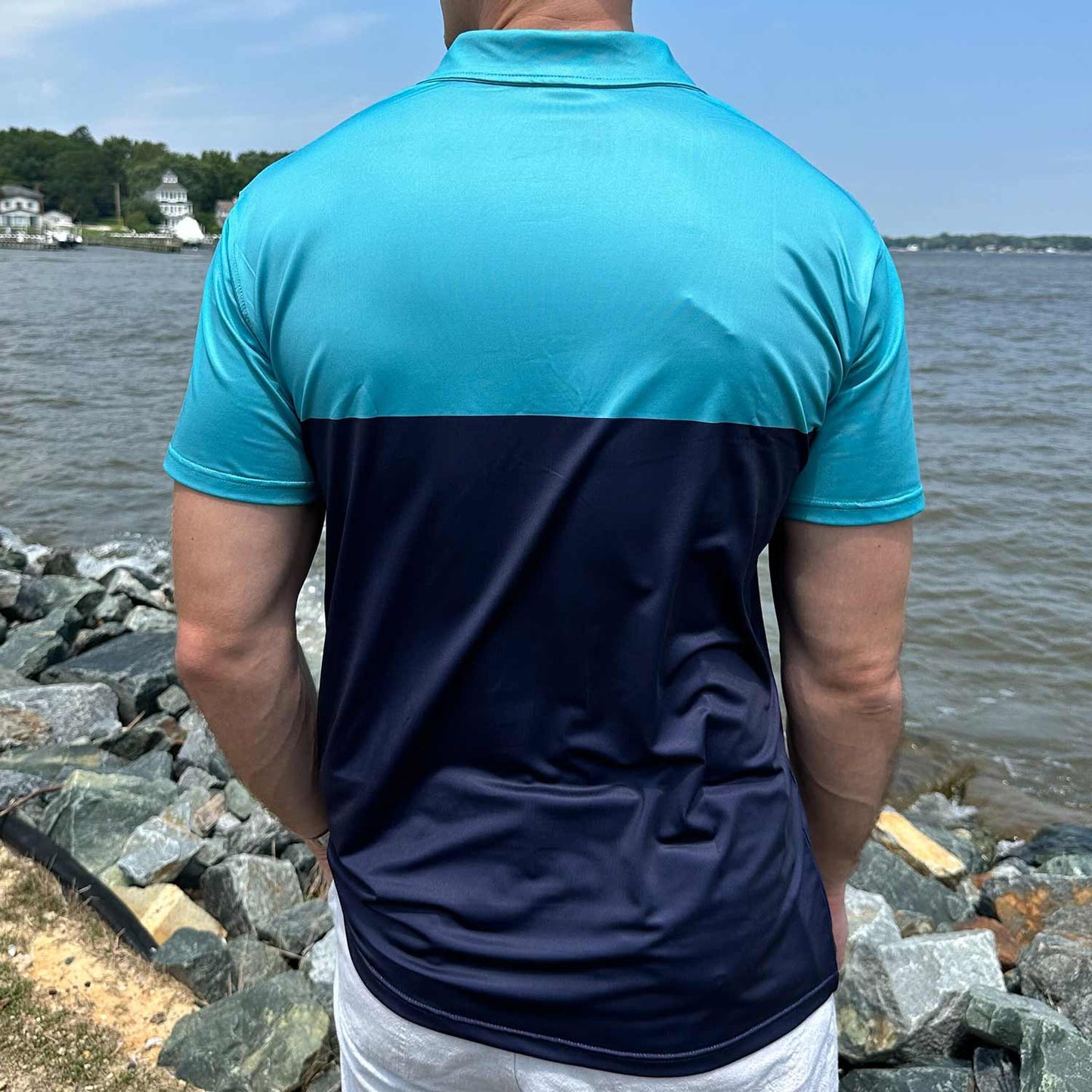 Rear view of man standing by coastal rocks and ocean wearing the Six Knots Into The Blue Performance Polo Shirt. This lightweight, moisture-wicking men's polo features bold robin's egg blue and navy stripes, crafted for UPF sun protection, breathability, and all-day comfort. Ideal for boating, saltwater fishing, beachside travel, and active coastal lifestyles.