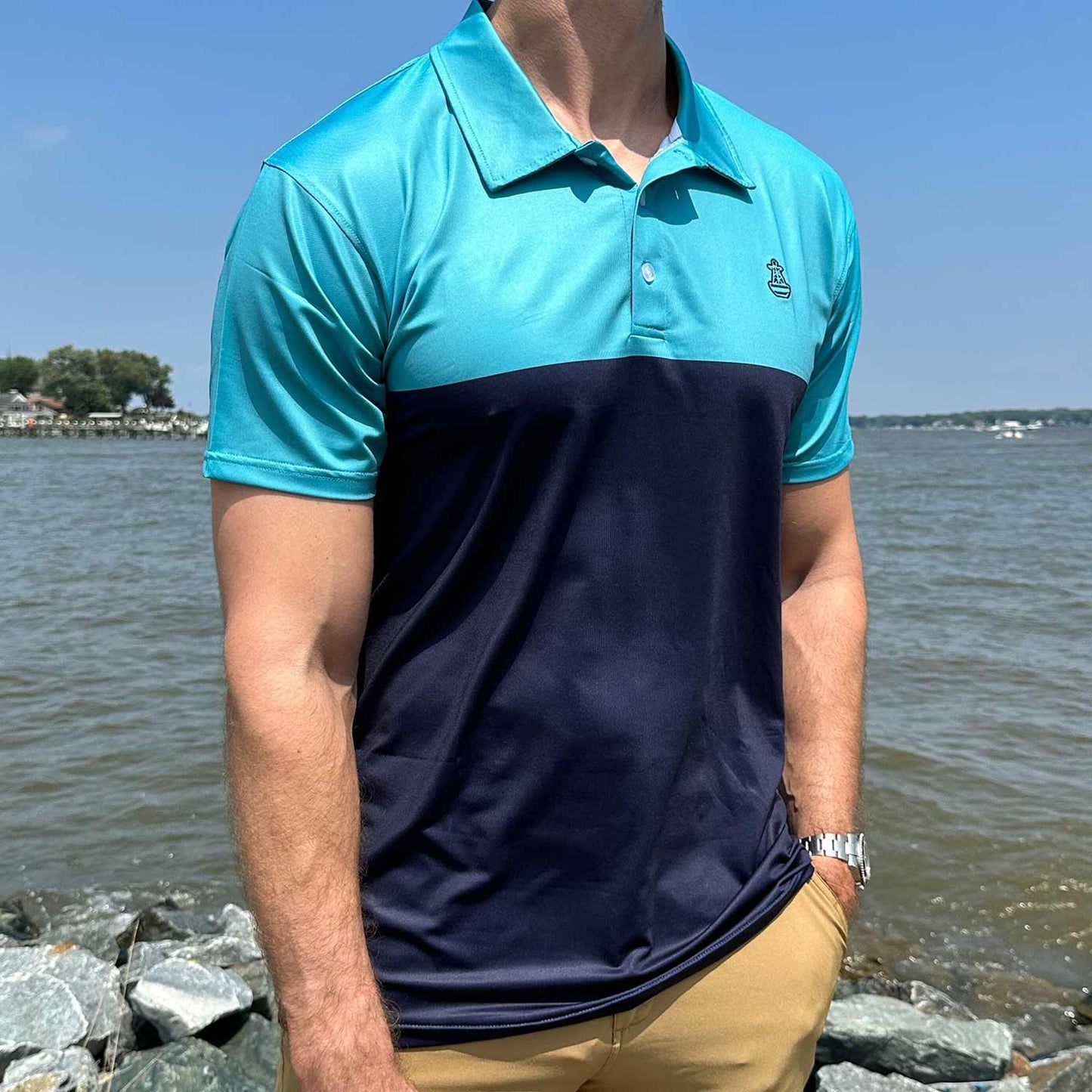 Man standing by coastal rocks and water wearing the Six Knots Into The Blue Performance Polo Shirt. This lightweight, moisture-wicking men's polo features bold robin's egg blue and navy stripes with the signature Six Knots buoy logo on the chest. Designed for sun protection and breathable comfort, perfect for boating, fishing, seaside travel, and warm-weather coastal living.