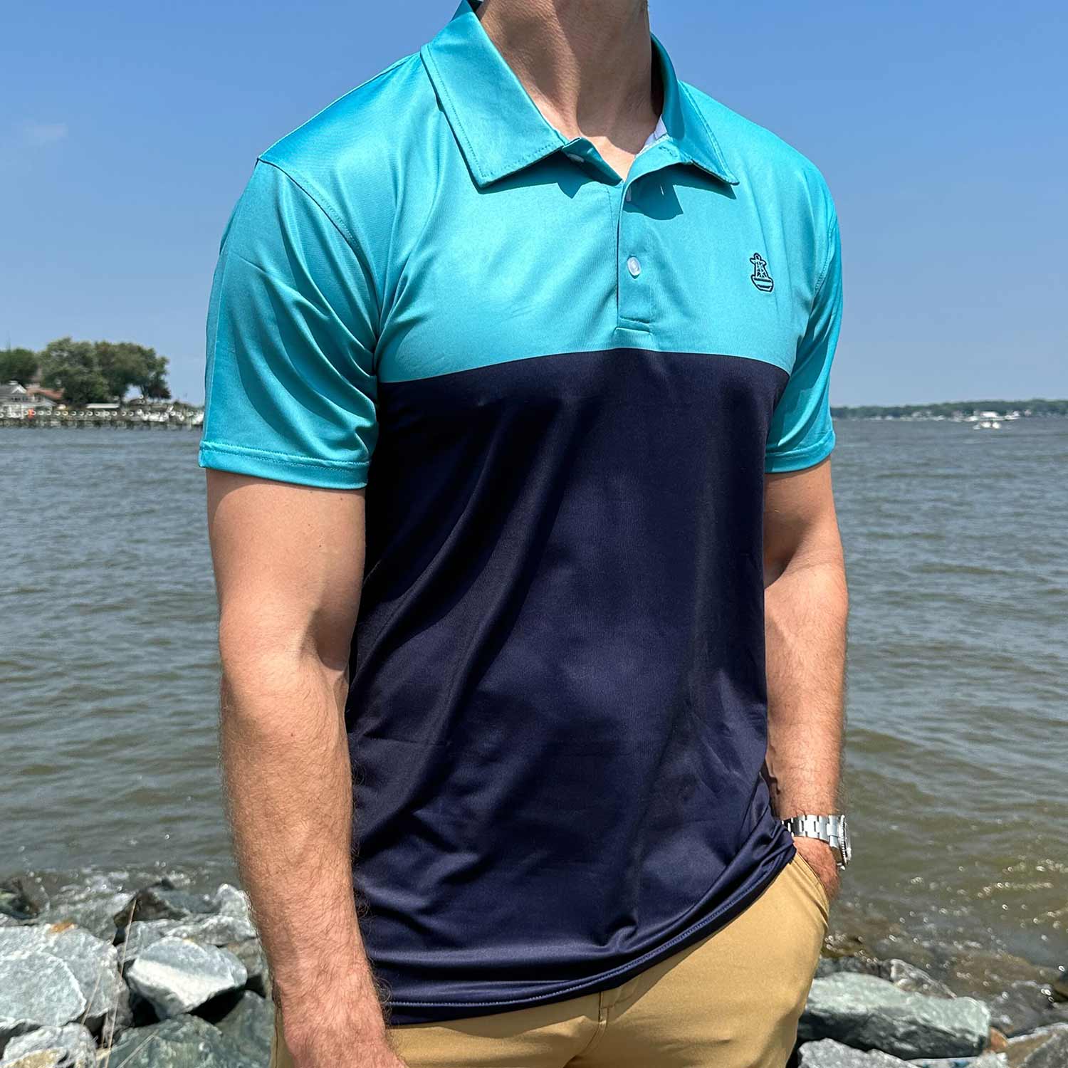 Man standing by coastal rocks and water wearing the Six Knots Into The Blue Performance Polo Shirt. This lightweight, moisture-wicking men's polo features bold robin's egg blue and navy stripes with the signature Six Knots buoy logo on the chest. Designed for sun protection and breathable comfort, perfect for boating, fishing, seaside travel, and warm-weather coastal living.