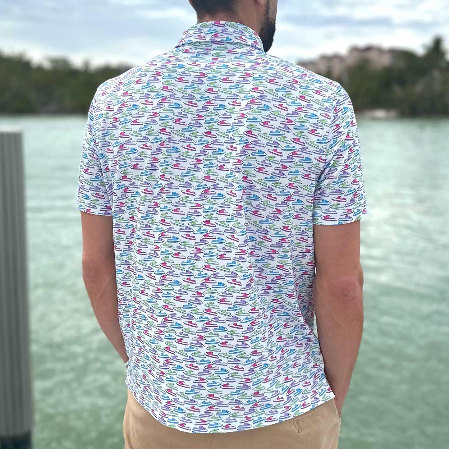 Rear view of a man standing on a dock in Florida wearing the Six Knots Just Send It Performance Polo Shirt. This lightweight, breathable men's polo features moisture-wicking fabric and a colorful all-over jet ski print in blue, green, pink, and purple. Perfect for boating, jet skiing, golfing, beach trips, and everyday coastal lifestyle activities.