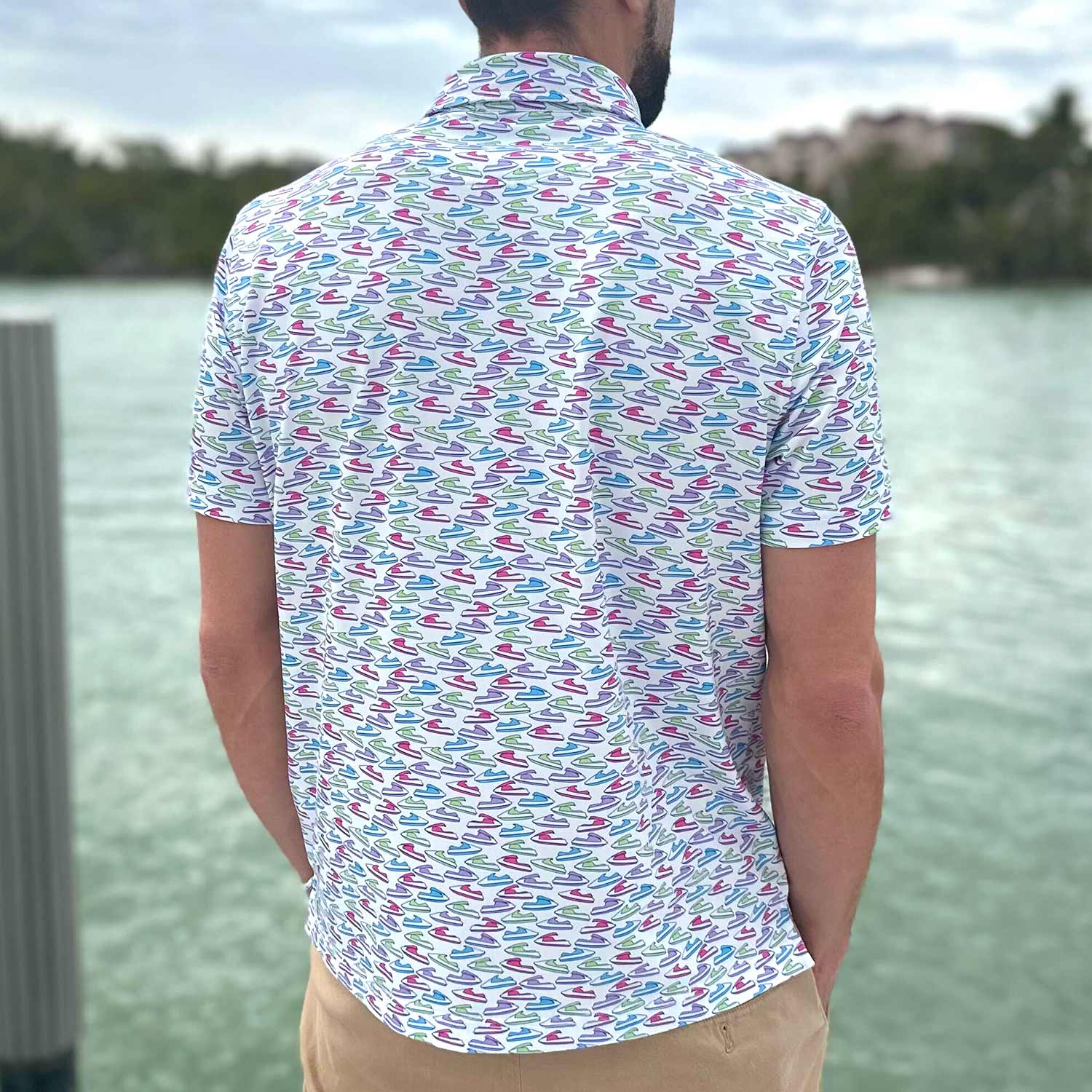 Rear view of a man standing on a dock in Florida wearing the Six Knots Just Send It Performance Polo Shirt. This lightweight, breathable men's polo features moisture-wicking fabric and a colorful all-over jet ski print in blue, green, pink, and purple. Perfect for boating, jet skiing, golfing, beach trips, and everyday coastal lifestyle activities.