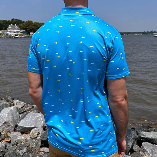 Rear view of man standing by coastal rocks and ocean water wearing the vibrant blue Six Knots Mahi Mahi Performance Polo Shirt. This lightweight polo for men features a bold white and yellow mahi mahi fish pattern, breathable moisture-wicking fabric, and UPF 50+ sun protection. Ideal for boating, beach days, golfing, and high-performance coastal wear.