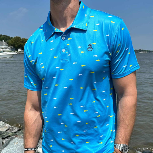 Man standing by coastal rocks and ocean water wearing the vibrant blue Six Knots Mahi Mahi Performance Polo Shirt. This lightweight, moisture-wicking men's polo features a bold white and yellow mahi mahi fish pattern, breathable fabric, and built-in UPF 50+ sun protection. Designed for all-day comfort and performance, it's perfect for boating, golfing, beach outings, and active coastal lifestyles.