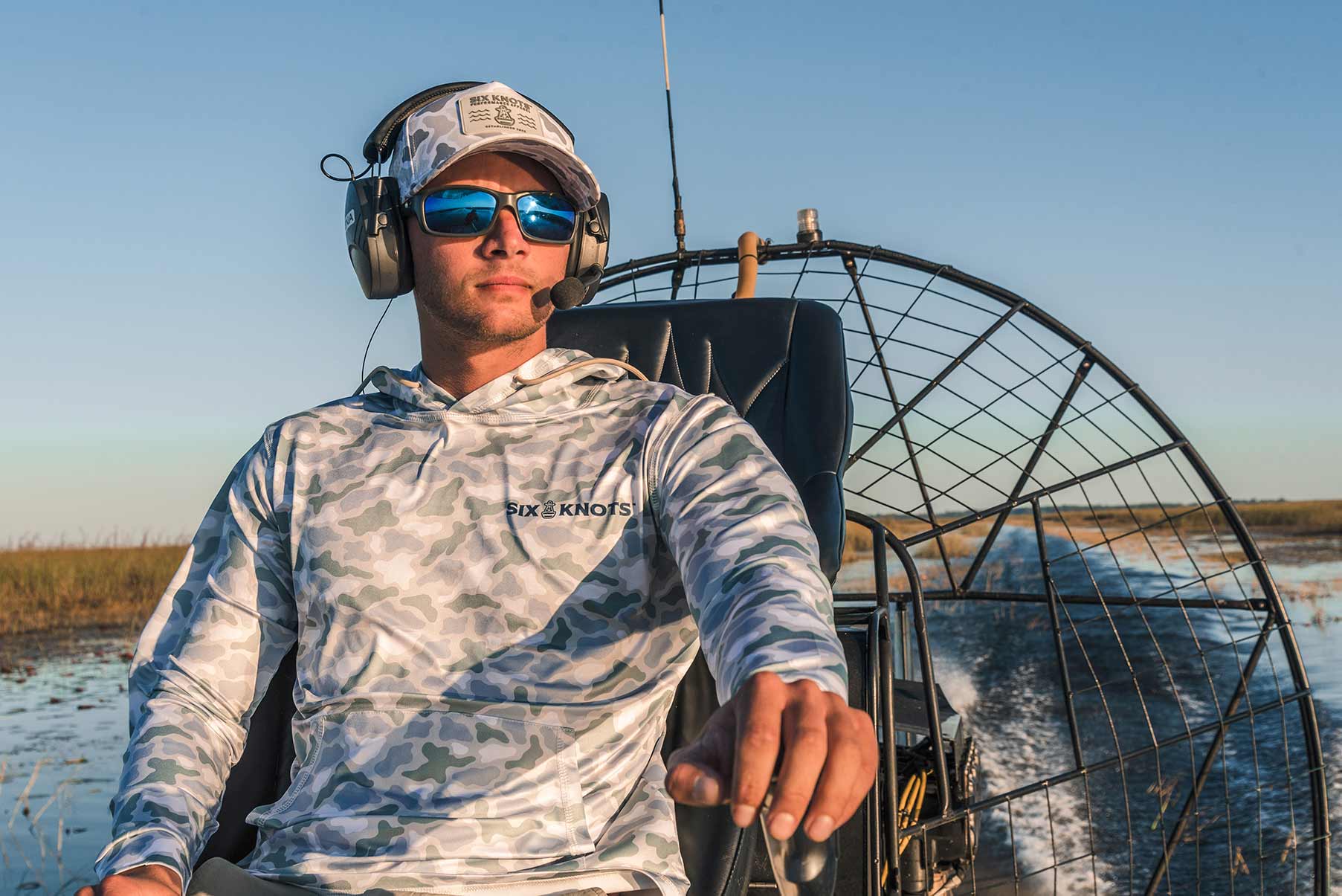 Man airboating in sunglasses, wearing the Six Knots water-resistant Coastal Camo Snapback Hat and Wetlands-pattern Chesapeake Performance Hoodie — lightweight, moisture-wicking boating apparel