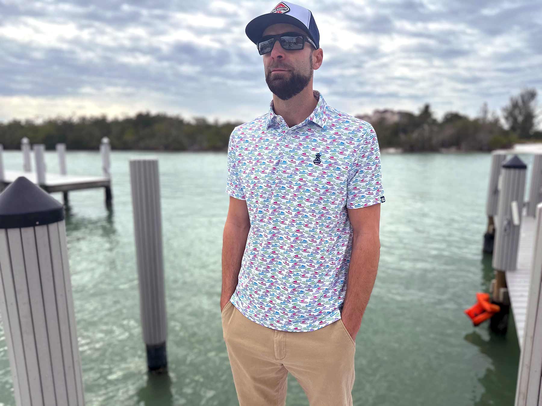 Man wearing sunglasses and a hat stands on a dock in Florida wearing the Six Knots Just Send It Performance Polo Shirt. This lightweight, breathable men's polo shirt features moisture-wicking fabric and a vibrant all-over jet ski print in blue, green, pink, and purple. Perfect for boating, jet skiing, golfing, beach outings, and everyday coastal lifestyle wear.