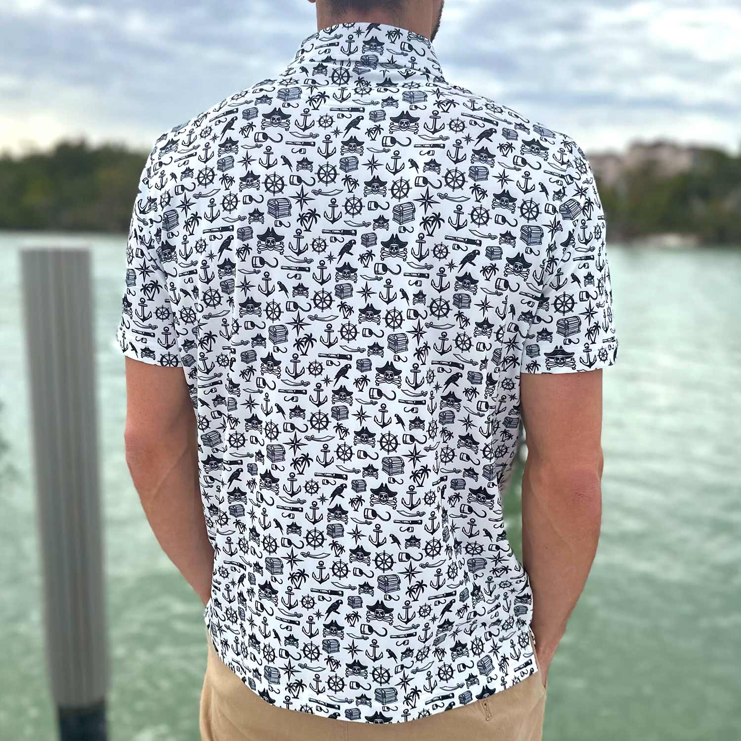 Rear view of a man standing on a dock in Florida wearing the Six Knots Pirate's Life Performance Polo Shirt. This breathable, lightweight men's polo features moisture-wicking fabric and a bold black and white nautical pirate print with treasure chests, skull and crossbones, cannons, hooks, and maritime symbols. Ideal for boating, golfing, beach outings, and coastal adventures.