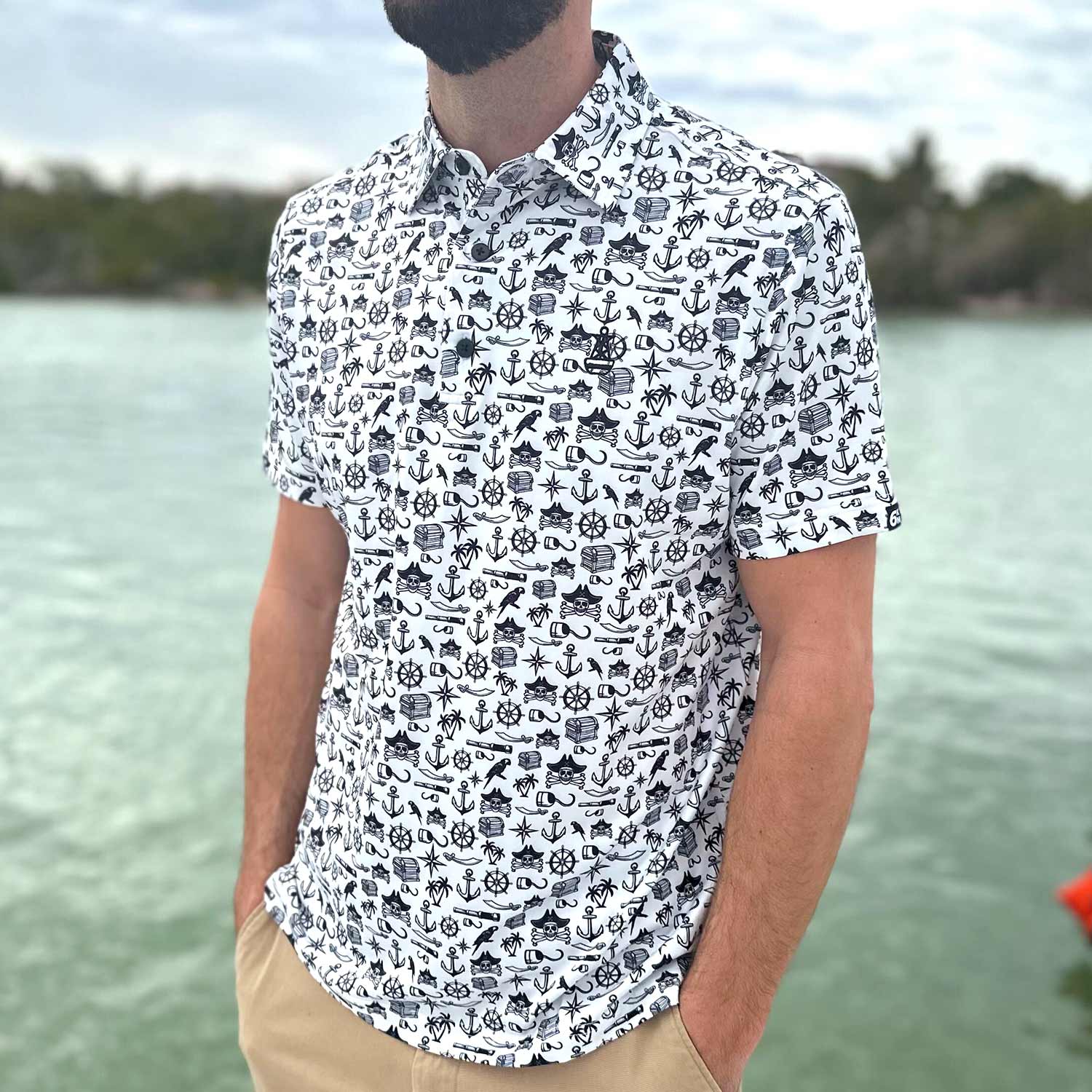 Man standing on a dock in Florida wearing the Six Knots Pirate's Life Performance Polo Shirt. This lightweight, moisture-wicking men's polo features an all-over black and white nautical pirate print with treasure chests, skull and crossbones, cannons, hooks, and maritime icons — perfect for boating, golfing, beach days, and coastal adventures